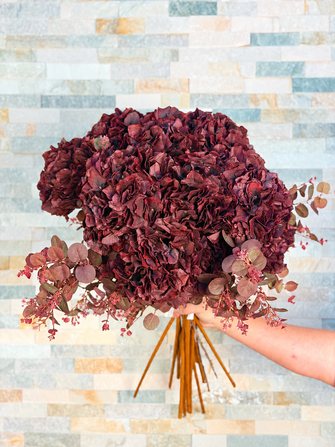 Fyllig bukett med konstgjord hortensia bordo i djupa vinröda nyanser, fotograferad framför en ljus stenvägg.
