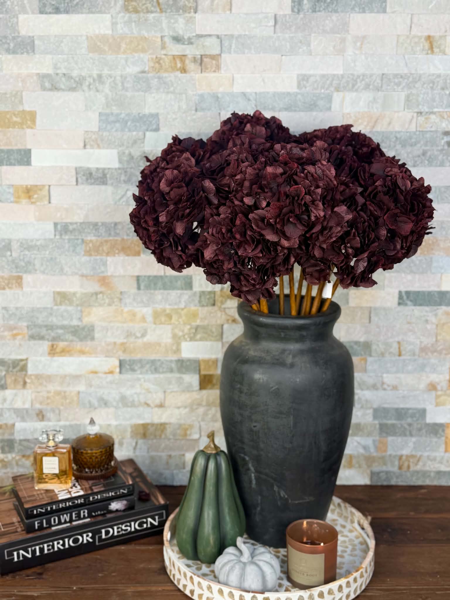 Konstgjord hortensia bordo 70 cm med fylliga vinröda blomklasar i en hög mörk vas på ett träbord framför en ljus stenvägg.